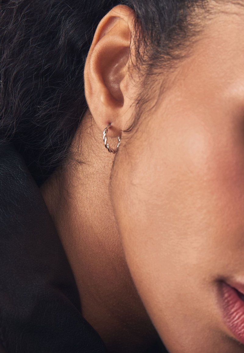 Silberner gedrehter Creolen-Ohrring, getragen an einem Nahaufnahme-Ohr, der ein minimalistisches Design mit glatter Textur vor dunklen, welligen Haaren zeigt.