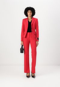 Un costume rouge en tissu lisse, comprenant un blazer cintré avec des accents noirs et un pantalon ajusté. Assorti d'un haut noir et d'un sac à main.
