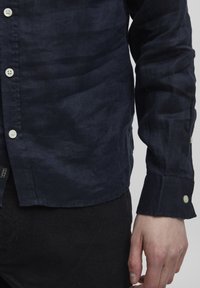 Camisa de lino azul oscuro con un acabado ligeramente texturizado, que presenta botones blancos y un puño estándar. Estilizada con un dobladillo redondeado.