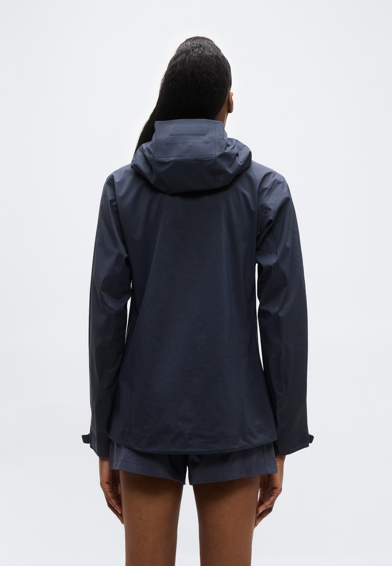 Personne portant une veste à capuche bleu marine et un short assorti, vue de dos sur un fond blanc uni.