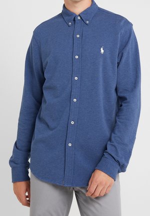 Mężczyzna ubrany w niebieską koszulę z długim rękawem i małym białym haftowanym logo na piersi, w połączeniu z jasnoszarymi spodniami.