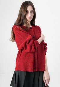 Cardigan en tricot rouge avec une finition texturée, présentant un col rond, des manches longues et des boutons noirs. Porté sur une jupe sombre.