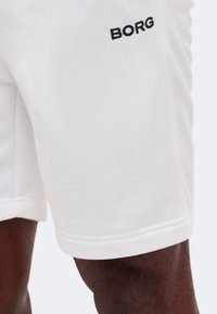 Κοντινό πλάνο στο μηρό ενός ατόμου με σκουρόχρωμο δέρμα που φοράει λευκά σορτς με μαύρο κείμενο "BORG" στο πάνω μπροστινό μέρος.