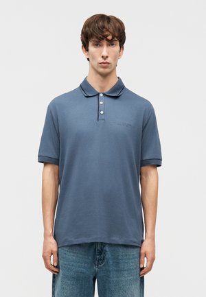 Junger Mann in blauem kurzärmligem Polohemd und blauer Jeans, der vor einem schlichten hellgrauen Hintergrund steht.