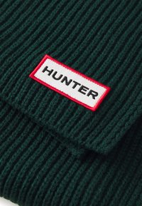 Tamnozelena pletiva s rebrastim uzorkom, s istaknutom bijelom etiketom obrubljenom crvenom, na kojoj velikim crnim slovima piše "HUNTER".