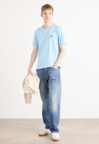 Ανοιχτό μπλε κοντομάνικο μπλουζάκι με λογότυπο "BOSS", συνδυασμένο με ξεθωριασμένα μπλε τζιν. Το μοντέλο κρατάει ένα μπεζ πουλόβερ και φοράει λευκά αθλητικά παπούτσια με μπεζ λεπτομέρειες.