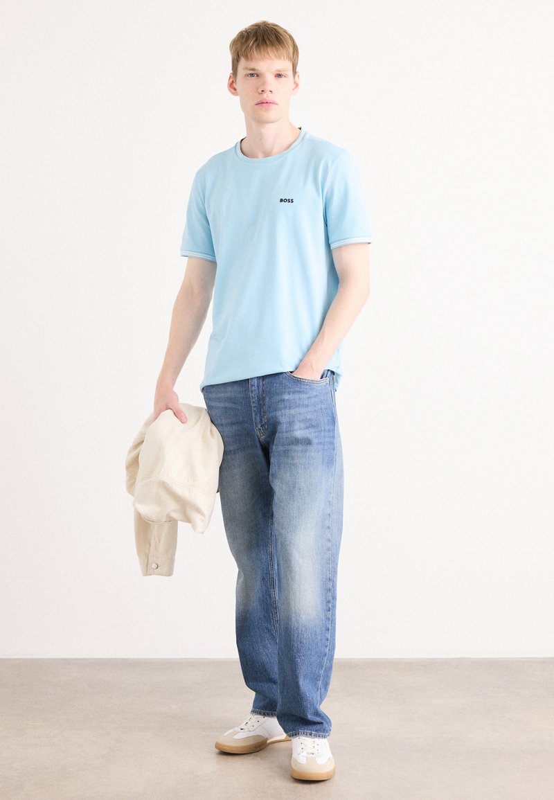 T-shirt bleu clair à manches courtes avec le logo "BOSS", associé à un jean bleu délavé. Le modèle tient un pull beige et porte des baskets blanches avec des accents beiges.