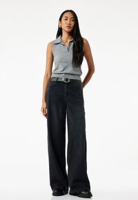Polo gris sin mangas, jeans negros de pierna ancha, ajustado con un cinturón gris que presenta una hebilla circular plateada, combinado con zapatos negros de suela gruesa.