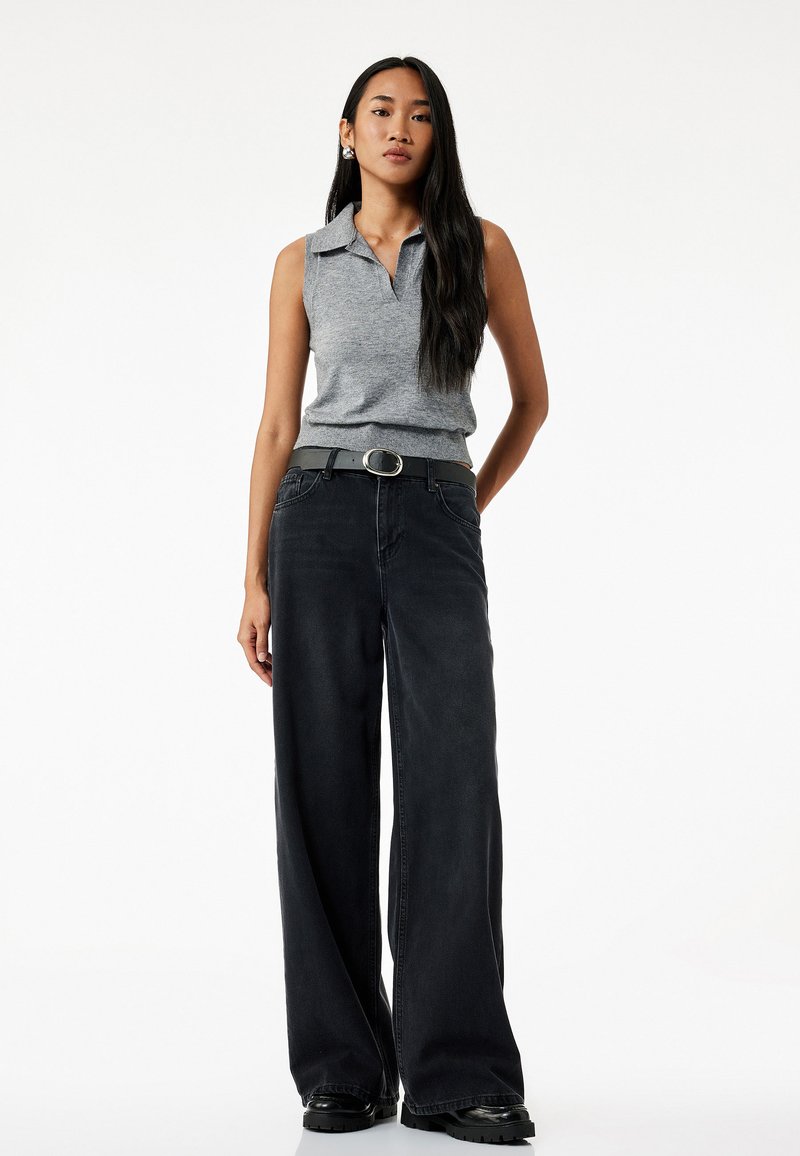 Polo gris sin mangas, jeans negros de pierna ancha, ajustado con un cinturón gris que presenta una hebilla circular plateada, combinado con zapatos negros de suela gruesa.