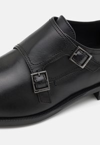 Black leather shoe with two silver buckles, smooth texture, rounded toe, and a low, slightly structured heel. Stitched detailing along edges.