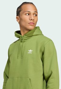 Žalias medvilnės džemperis su priekinėmis kišenėmis, su raišteliu reguliuojama kapuce ir balta Adidas logotipu ant krūtinės. Lygi tekstūra ir laisvas kirpimas.