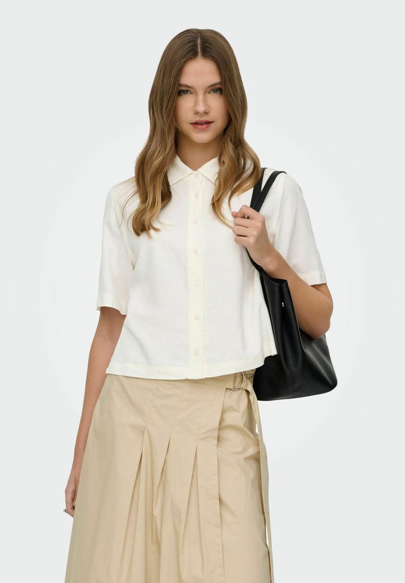 Jeune femme aux longs cheveux portant une chemise blanche à manches courtes boutonnée et une jupe plissée beige, tenant un sac à bandoulière noir.