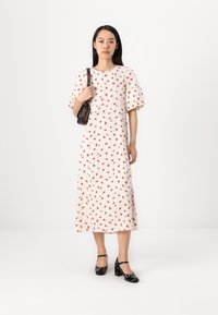 Witte bloemenjurk met rode rozenpatronen, korte pofmouwen en een jurk van gemiddelde lengte. Model houdt een kleine bruine tas vast. Zwarte hoge hakken.