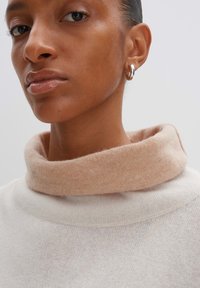 Hellbeiger Pullover mit einem weichen, gerollten pfirsichfarbenen Kragen. Die Textur wirkt gestrickt, mit subtilen Details im Halsbereich.