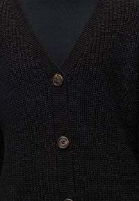 Gros plan sur le devant d’un cardigan en maille noire avec de gros boutons marron et un col en V sur un haut noir.