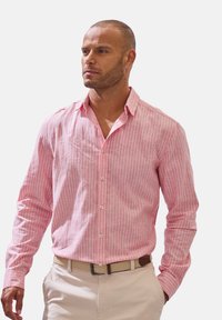 Hemd aus hellrosa gestreifter Baumwolle mit langen Ärmeln, Button-Down-Kragen und weißen Knöpfen. Getragen mit einem beigen Gürtel und hellen Hosen.