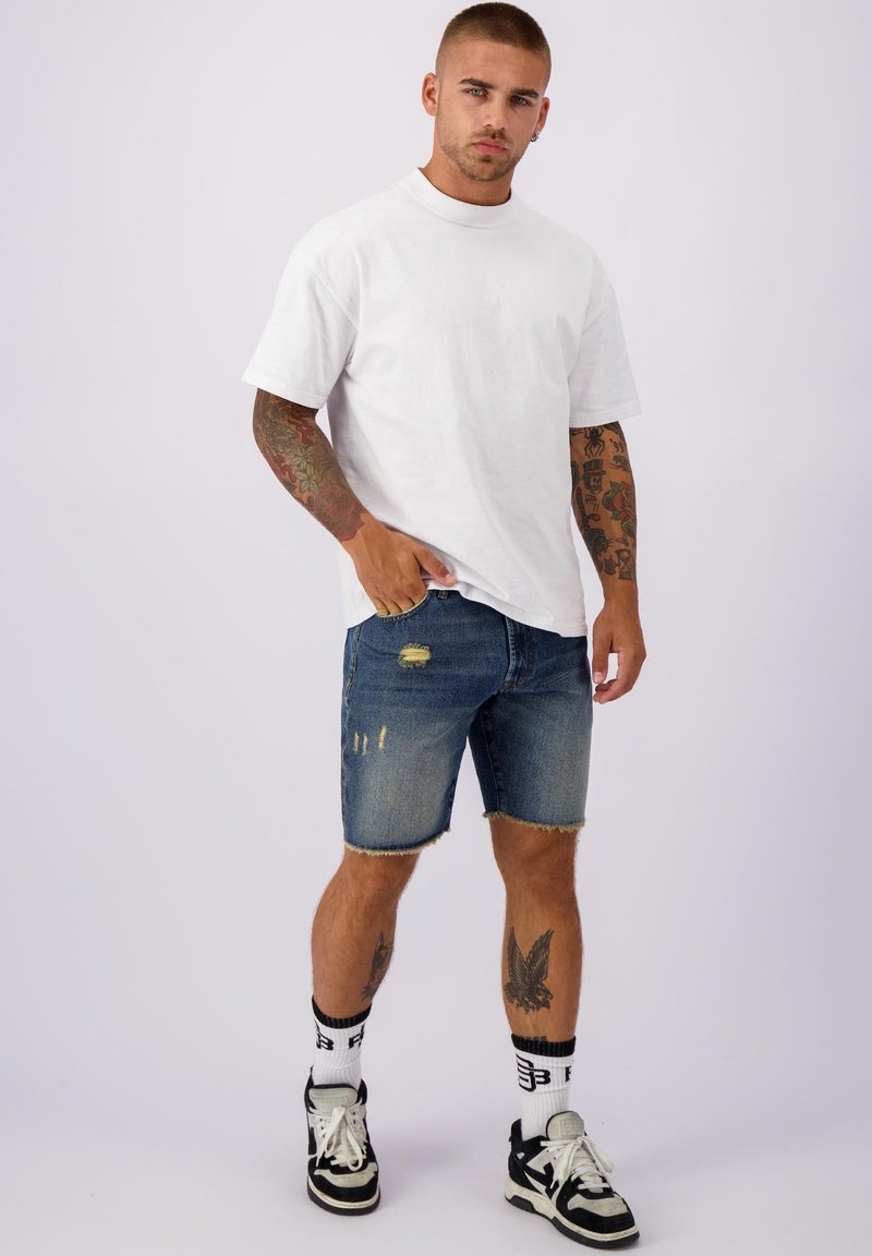 Een man staat zelfverzekerd, gekleed in een wit T-shirt, versleten denim shorts en zwarte sneakers, terwijl hij zijn kleurrijke tatoeages toont.