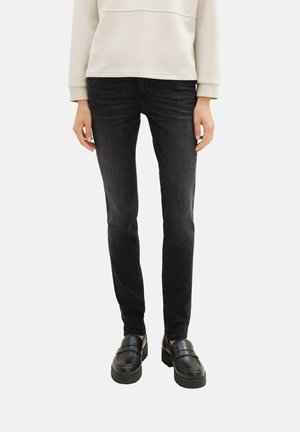 Person iført tætsiddende sorte jeans, cremefarvet sweatshirt og sorte chunky loafers, stående mod en ensartet lys baggrund.