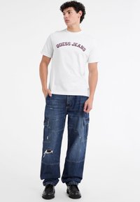 Balta medvilninė marškinė su "GUESS JEANS" tamsiai raudonais užrašais, derinami su mėlynomis džinsinėmis cargo kelnėmis, kurios turi kiaurai dėvėtų vietų ir dideles kišenes.