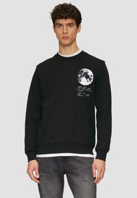 Zwarte sweatshirt met maangrafiek en tekst op de linkerborst. Ronde hals en geribbelde manchetten. Draagt over een wit shirt en in combinatie met jeans.