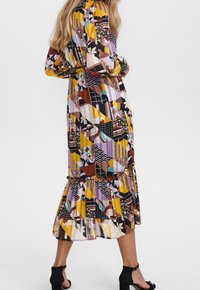 Robe longue multicolore à manches longues, avec des motifs géométriques et floraux dans des nuances de jaune, noir, violet et marron. Ourlet évasé.