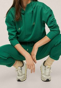Groene hoodie met een zak aan de voorkant en koorden, gecombineerd met bijpassende groene broek. Bevat beige sneakers met grijze accenten en geribbelde sokken.