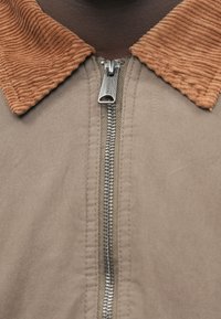 Μπεζ μπουφάν με ασημένιο φερμουάρ και καστανό κεραμιδί γιακά, κοντινό πλάνο που δείχνει το φερμουάρ με ετικέτα "premium".