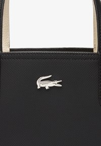 Sac à main noir texturé avec un logo crocodile argenté centré sous la poignée, montrant la partie supérieure du sac.