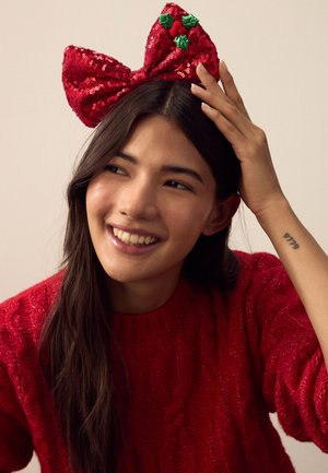 Red sequin bow headband with green holly accents. Model wears a red knit sweater. Hair is dark, styled gently, with a soft, textured appearance.