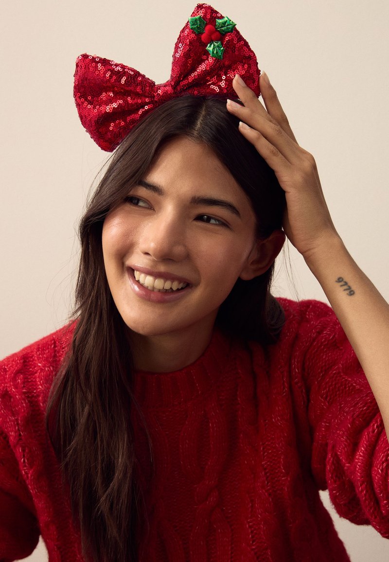 Rotes Haarband mit Pailletten-Schleife und grünen Stechpalmenakzenten. Das Modell trägt einen roten Strickpullover. Das Haar ist dunkel, sanft gestylt und hat ein weiches, strukturiertes Aussehen.