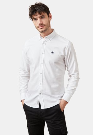 Camisa blanca de botones con cuello puntiagudo, que presenta un pequeño logo en color marino en el pecho. Mangas largas con puños de botones y textura de tela visible.