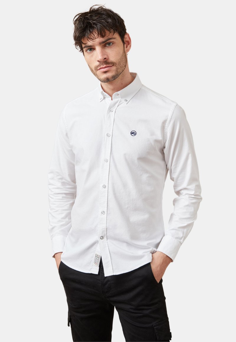 Camisa blanca de botones con cuello puntiagudo, que presenta un pequeño logo en color marino en el pecho. Mangas largas con puños de botones y textura de tela visible.