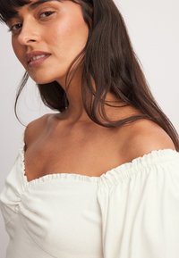 Biela blúzka s odhalenými ramenami, riaseným výstrihom a dlhými, nadýchanými rukávmi. Materiál vyzerá mäkko s textúrovaným povrchom.