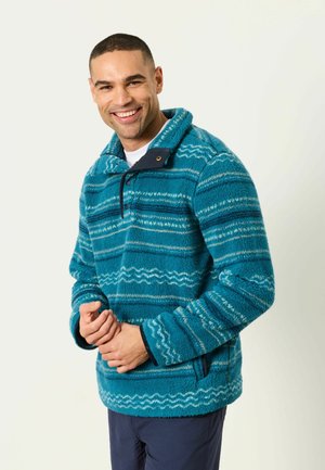 Lächelnder Mann, der einen blau gemusterten Fleecepullover mit halbgeknöpftem Kragen und eine marineblaue Hose trägt, steht vor einem schlichten hellen Hintergrund.
