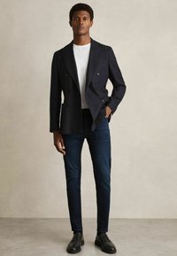 Mornarica dvostruko kopčana blazer, bijela majica, tamni traperice i crne cipele s vezicama. Glatka tkanina, kroj prilagođen tijelu, moderni stil.