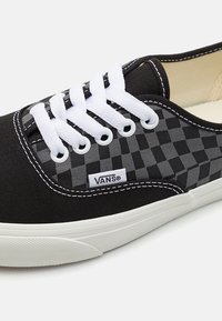 Baskets Vans à carreaux noirs et gris avec lacets blancs, coutures blanches et semelle en caoutchouc blanche, inclinées pour montrer les détails latéraux et avant.