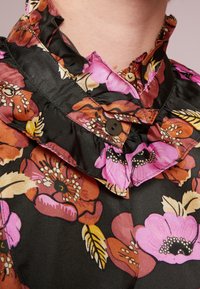 Blusa nera floreale con grandi fiori rosa, arancioni e gialli, colletto ondulato e chiusura con bottone marrone. Tessuto di trama liscia.