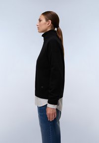 Musta poolapainen villapaita, jossa on rento malli, ribbikuvioiset hihansuut ja lyhyempi pituus, kerroksittain vaalean raidallisen paidan päällä. Farkut täydentävät asukokonaisuuden.