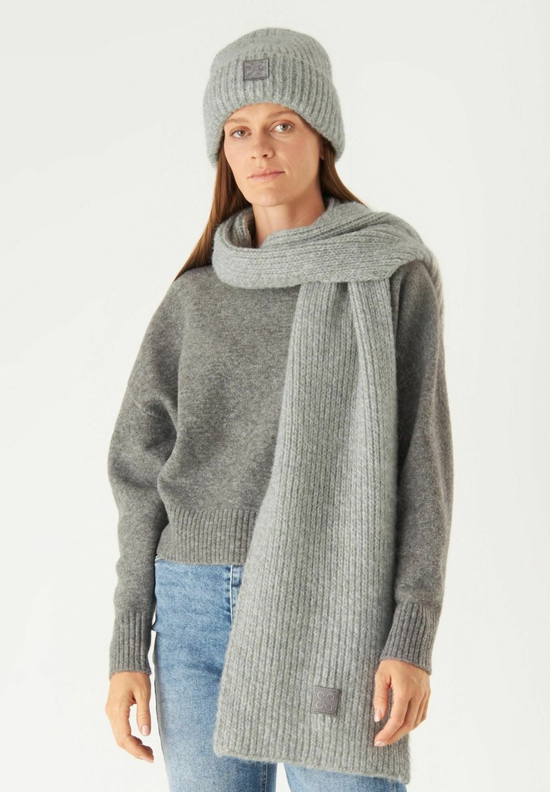 Bonnet en tricot gris et écharpe côtelée en matériau doux, portés avec un pull gris et un jean bleu. Comprend un petit logo sur l'écharpe.