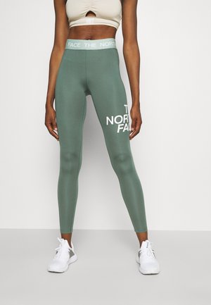Personne portant des leggings taille haute vert sauge avec le logo "The North Face" sur la cuisse droite et des baskets blanches, debout devant un fond uni.