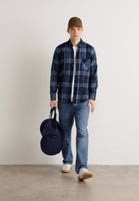 Camisa xadrez em tons de azul marinho, jeans azuis e ténis brancos. A carregar uma mala de viagem azul marinho com alças pretas. Fundo simples.