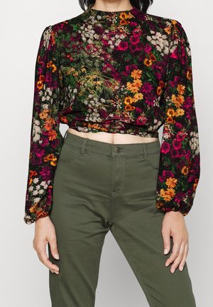 Vrouw draagt een bloemtop met lange mouwen en een hoge hals, bedrukt met oranje, paarse en witte bloemen, gecombineerd met olijfgroene broek met hoge taille.