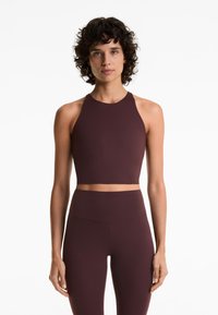 Žena s kovrčavom tamnom kosom nosi bezrukavi tamnosmeđi crop top i odgovarajuće tajice visokog struka, stojeći uz bijelu pozadinu.