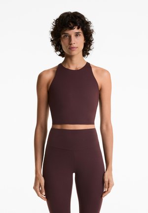 Žena s kovrčavom tamnom kosom nosi bezrukavi tamnosmeđi crop top i odgovarajuće tajice visokog struka, stojeći uz bijelu pozadinu.