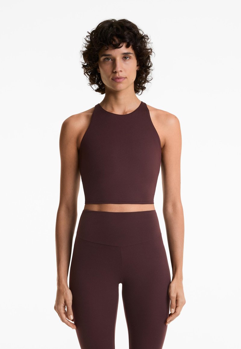 Žena s kovrčavom tamnom kosom nosi bezrukavi tamnosmeđi crop top i odgovarajuće tajice visokog struka, stojeći uz bijelu pozadinu.