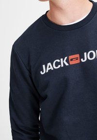 Uomo che indossa una felpa blu navy con il logo "JACK & JONES" stampato in bianco e rosso sul petto.