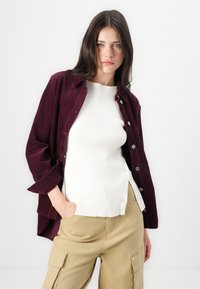Bordeauxkleurig corduroy overhemd met knopen over een wit geribd topje, gecombineerd met lichtbeige cargobroeken met zijzakken.