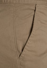 Beige stof met een gladde textuur, met een zijzak en gedetailleerde stiksels. Zichtbare naden en een klassiek ontwerp.