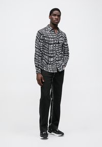 Svartvit skjorta med knappar och ett upprepande textmönster; ihop med svarta byxor med en kontrasterande vit söm; svarta sneakers.