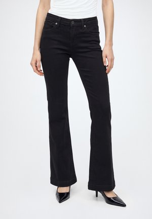 Femme portant un jean bootcut noir et des talons hauts pointus noirs, debout, les mains détendues le long du corps sur fond blanc.